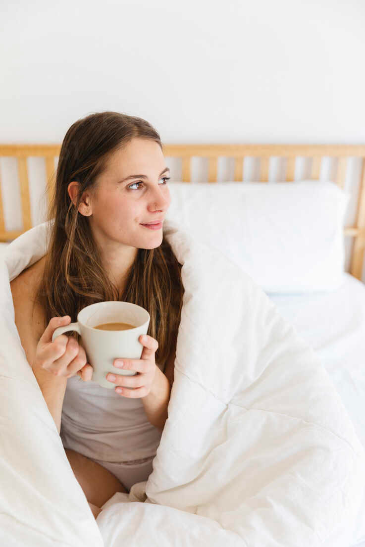 young-woman-holding-coffee-cup-while-wrapped-in-white-blanket-at-home-MRAF00692.png